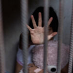 Child behind bars, hand raised, holding a teddy bear.