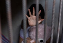 Child behind bars, hand raised, holding a teddy bear.