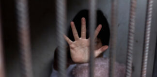 Child behind bars, hand raised, holding a teddy bear.