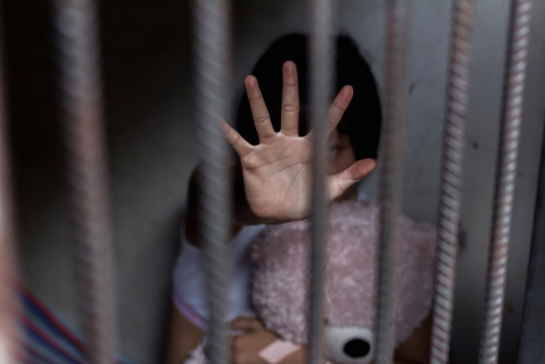 Child behind bars, hand raised, holding a teddy bear.