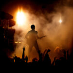 Guitarist performing on stage with audience and dramatic lighting.