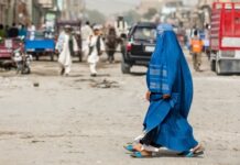 Woman in blue burqa walking through busy street scene.