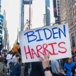 Person holding Biden Harris sign in crowded city street.