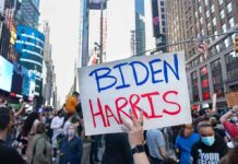 Person holding Biden Harris sign in crowded city street.