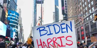 Person holding Biden Harris sign in crowded city street.