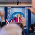 Press secretary at podium, answering reporters questions, raised hands.