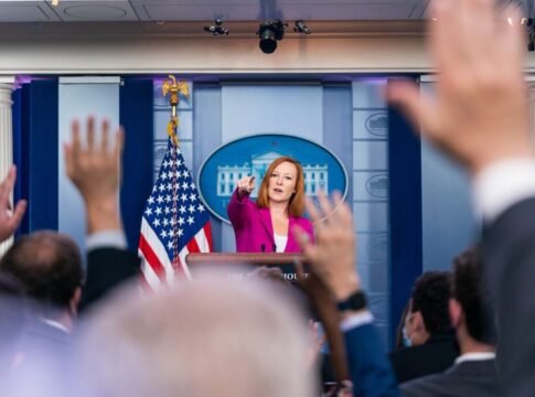 Press secretary at podium, answering reporters questions, raised hands.