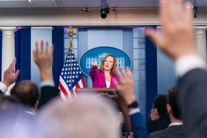 Press secretary at podium, answering reporters questions, raised hands.