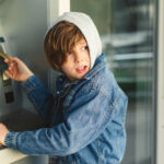 Child using an ATM machine outdoors, wearing denim jacket.