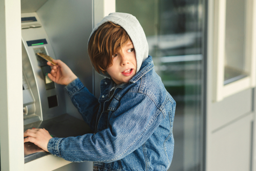 Child using an ATM machine outdoors, wearing denim jacket.