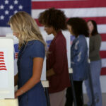 People voting in booths with U.S. flags in background.