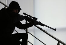 Person in dark clothing aiming a rifle indoors.