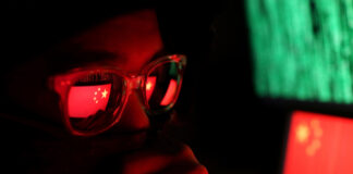 Person wearing glasses with a reflection of the Chinese flag, surrounded by computer screens