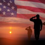 Silhouetted soldiers saluting against an American flag backdrop during sunset