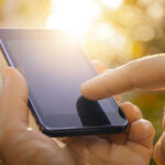 Finger touching smartphone screen with sunlight background.