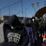 People in hoodies and masks walking beside a fence.