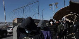 People in hoodies and masks walking beside a fence.