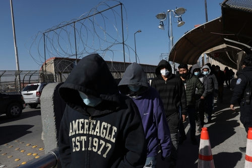 shutterstock_2089375438 People in hoodies and masks walking beside a fence.