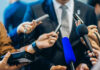 Final Report Pending—Speculation Erupts Journalists holding microphones interview a person in a suit.