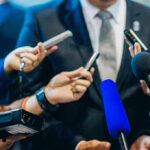 Journalists holding microphones interview a person in a suit.
