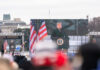 A large screen displays a speaker at a political rally with American flags in the foreground