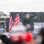 A large screen displays a speaker at a political rally with American flags in the foreground