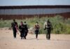 Group walking on a dusty path near a tall border fence