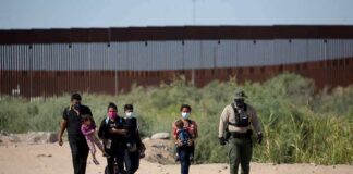 Group walking on a dusty path near a tall border fence