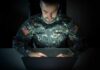 Deepfake Vets Hustle Patriot Cash Soldier in camouflage uniform using a laptop in a dark room