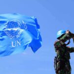 UN peacekeeper with blue helmet looks through binoculars beside UN flag