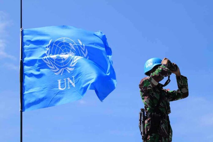 shutterstock_1480711058-1 UN peacekeeper with blue helmet looks through binoculars beside UN flag