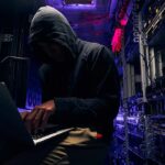 Hooded person using a laptop in a dark server room