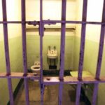 Interior of a prison cell with a toilet, sink, and metal bars