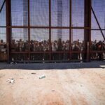 Border Chaos Pushes City Into Emergency Crowd stands behind tall metal fence at border crossing