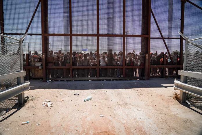 shutterstock_2298883197 Crowd stands behind tall metal fence at border crossing
