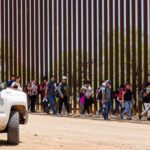 Group of people walking along a tall border fence beside a patrol vehicle