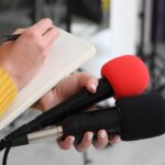 King’s Cold Downgrade Stuns Sussexes Journalist holding two microphones while writing in a notebook