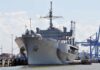 Large gray naval ship docked at an industrial port