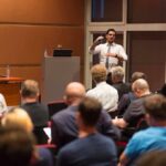 Taylor Cheers, Room Winces — What Happened? Speaker addresses audience in a conference room
