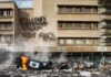 Riot Inferno – Macron Vows Retribution Burning overturned police car in front of a damaged building