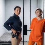 Police officer stands beside inmate in orange uniform inside a jail cell