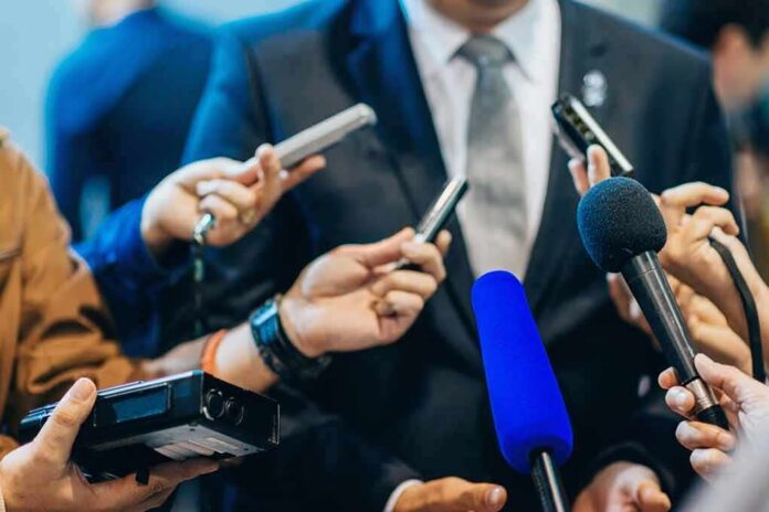 shutterstock_471987565 Man in suit surrounded by microphones at press conference