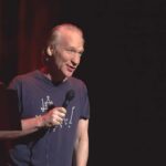 Bill Maher performing stand-up with a microphone on stage